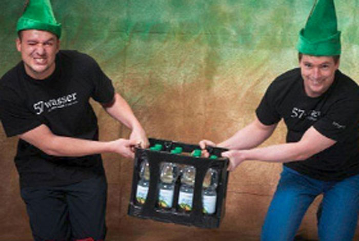 Die Gründer von 57wasser tragen grüne Hüte und halten einen Kasten des regionalen Mineralwassers zwischen sich