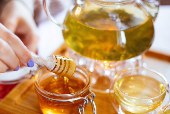 Hand mit Honiglöffel über einem Honigglas mit weißem Tee im Hintergrund. Der Honig stammt aus lokaler Erzeugung in Siegen.